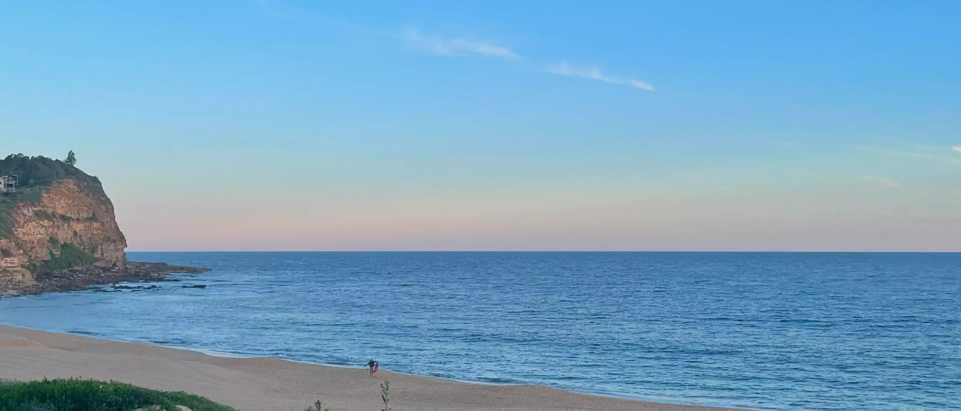 Beach scene with a person walking along the shore, blue ocean, and clear sky.
