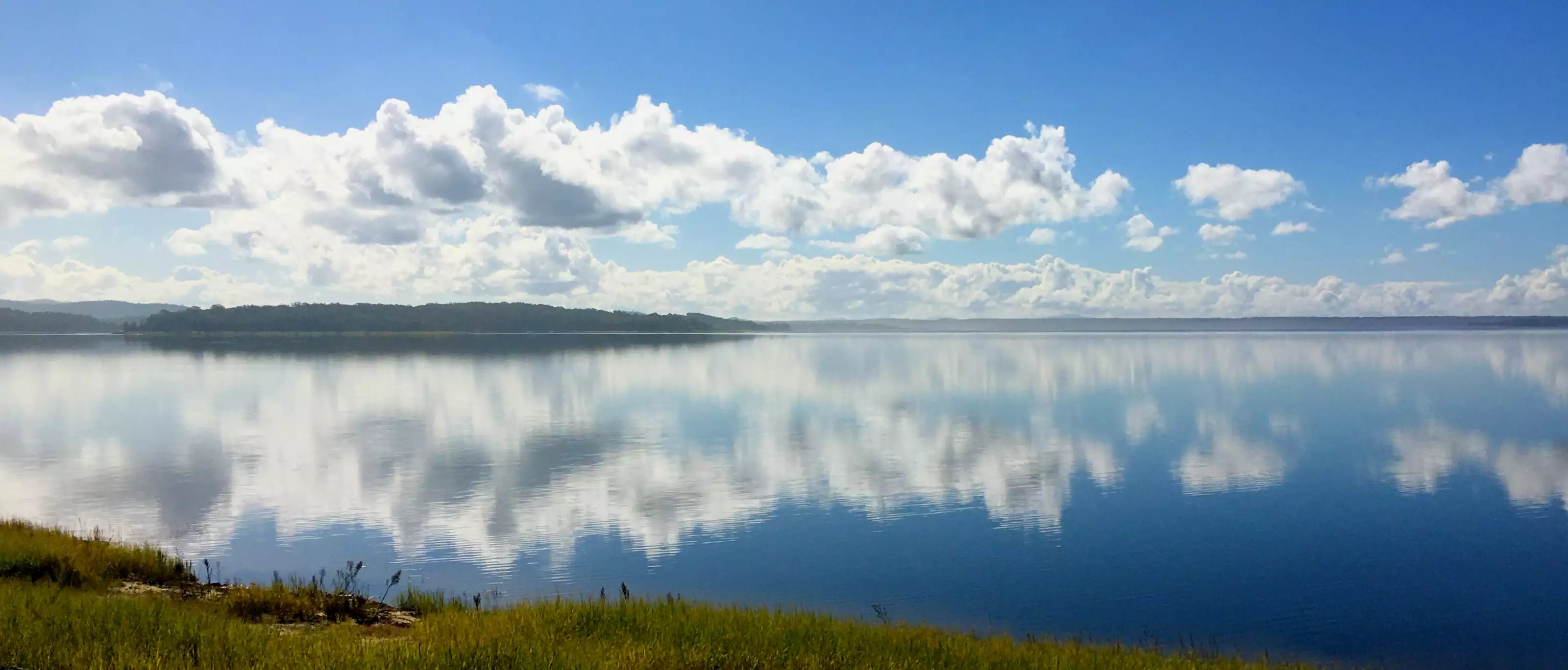 Panoramic view of a lake with clouds reflected on the water under a blue sky.